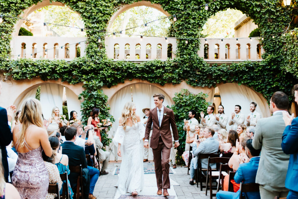 A couple walking down the aisle hand in hand after their ceremony at Tlaquepaque, one of the best wedding venues in Sedona. Guests are around them, clapping.