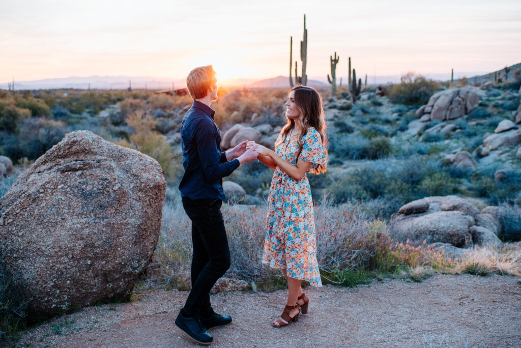 A couple during their engagement photo session in Phoenix, holding hands in front of the sun setting behind tall cacti.